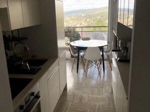 A narrow kitchen with a dining area, a sink, and a view of a town through large windows. - Akwaba 1-Bedroom Apartment with Ocean View, Terrace, and Pool (Cassis)