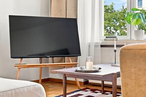 A room with a flat-screen TV on a stand, a small table with a candle, a potted plant, and a window with a view of trees. - Timeless Nordic | Quiet Street | Parquet | Lift (Helsinki)
