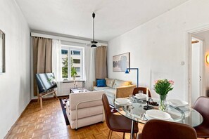 A modern living room with a glass dining table set for two, a sofa, a coffee table, and a painting on the wall. - Timeless Nordic | Quiet Street | Parquet | Lift (Helsinki)