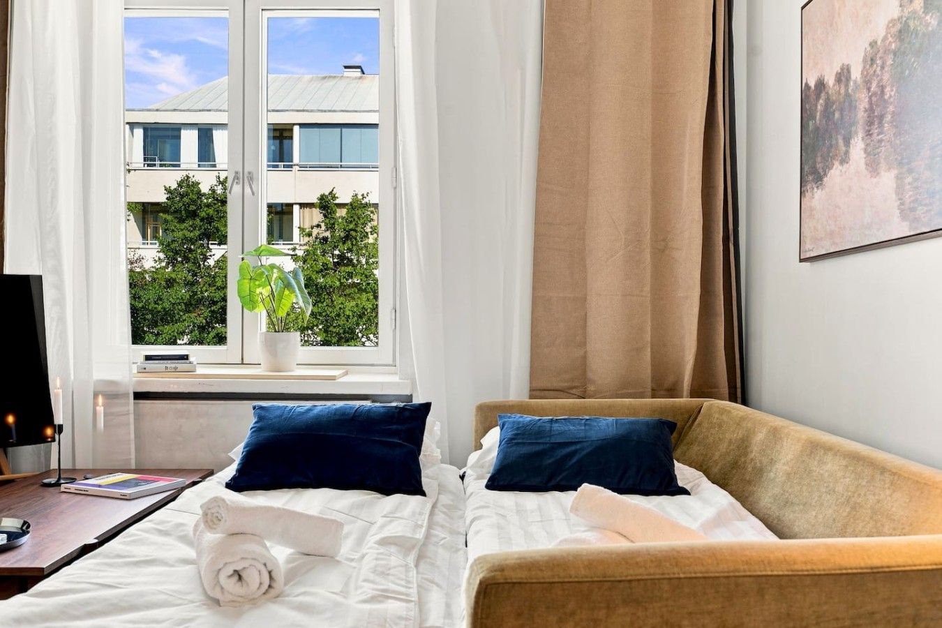 A hotel room with a bed, a desk with a laptop, a window with a view of buildings and trees, and a painting on the wall.