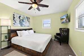 A bedroom with a bed, a ceiling fan, a window, a chair, and a painting on the wall. - Dave’s Place (Bentonville)
