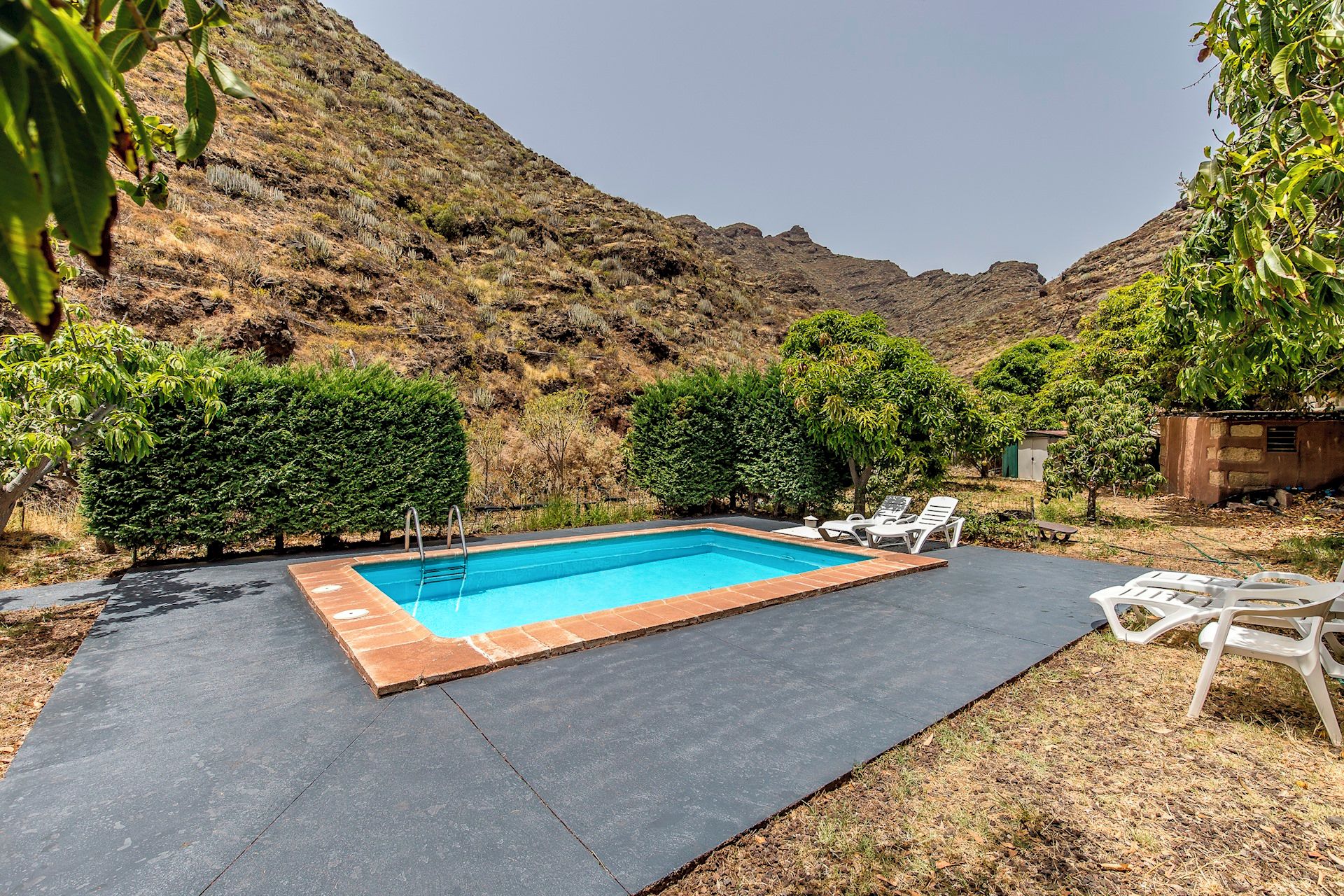 Une piscine rectangulaire dont les bords sont recouverts de carreaux, entourée d’une terrasse dallée. On aperçoit, au fond, des chaises longues blanches et un petit abri de jardin.