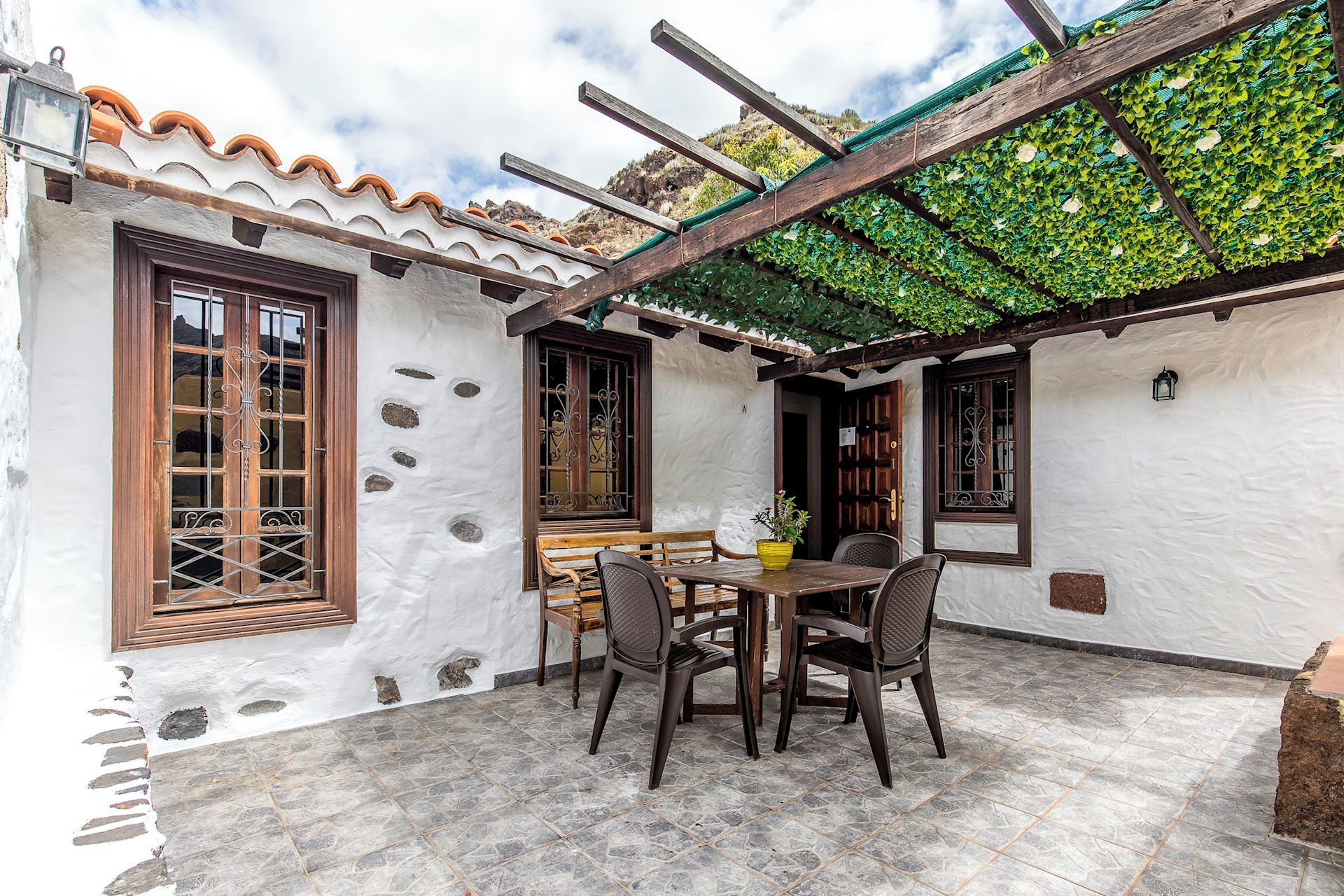 Une terrasse avec une table et des chaises, une pergola couverte de plantes grimpantes et un mur blanc agrémenté de cadres décoratifs pour les fenêtres.