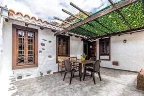 Een terras met een tafel en stoelen, een pergola begroeid met klimplanten, en een witte muur met decoratieve raamkozijnen.