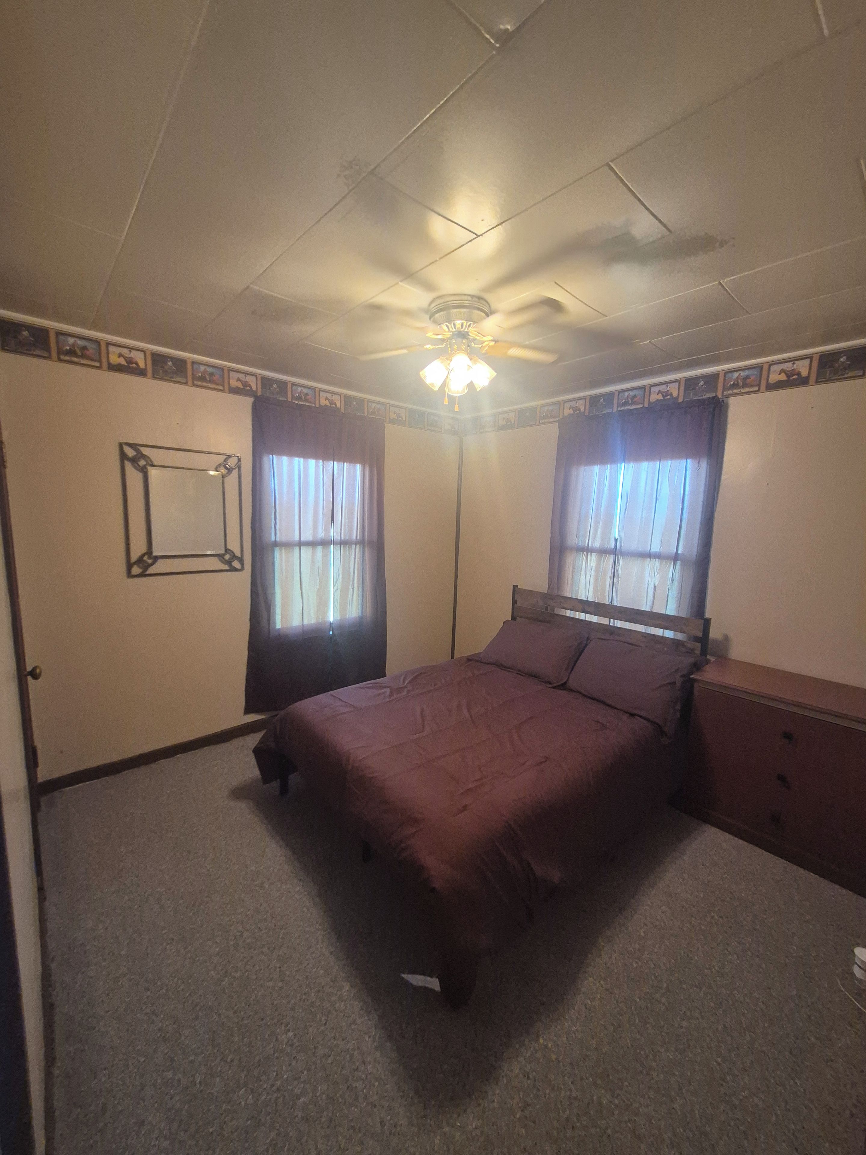 Ein Schlafzimmer mit einem Bett, einem Schminktisch, einem Deckenventilator und zwei mit Vorhängen versehenen Fenstern.