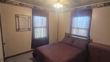 Ein Schlafzimmer mit einem Bett, einem Schminktisch, einem Deckenventilator und zwei mit Vorhängen versehenen Fenstern.