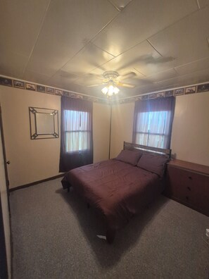 Ein Schlafzimmer mit einem Bett, einem Schminktisch, einem Deckenventilator und zwei mit Vorhängen versehenen Fenstern.