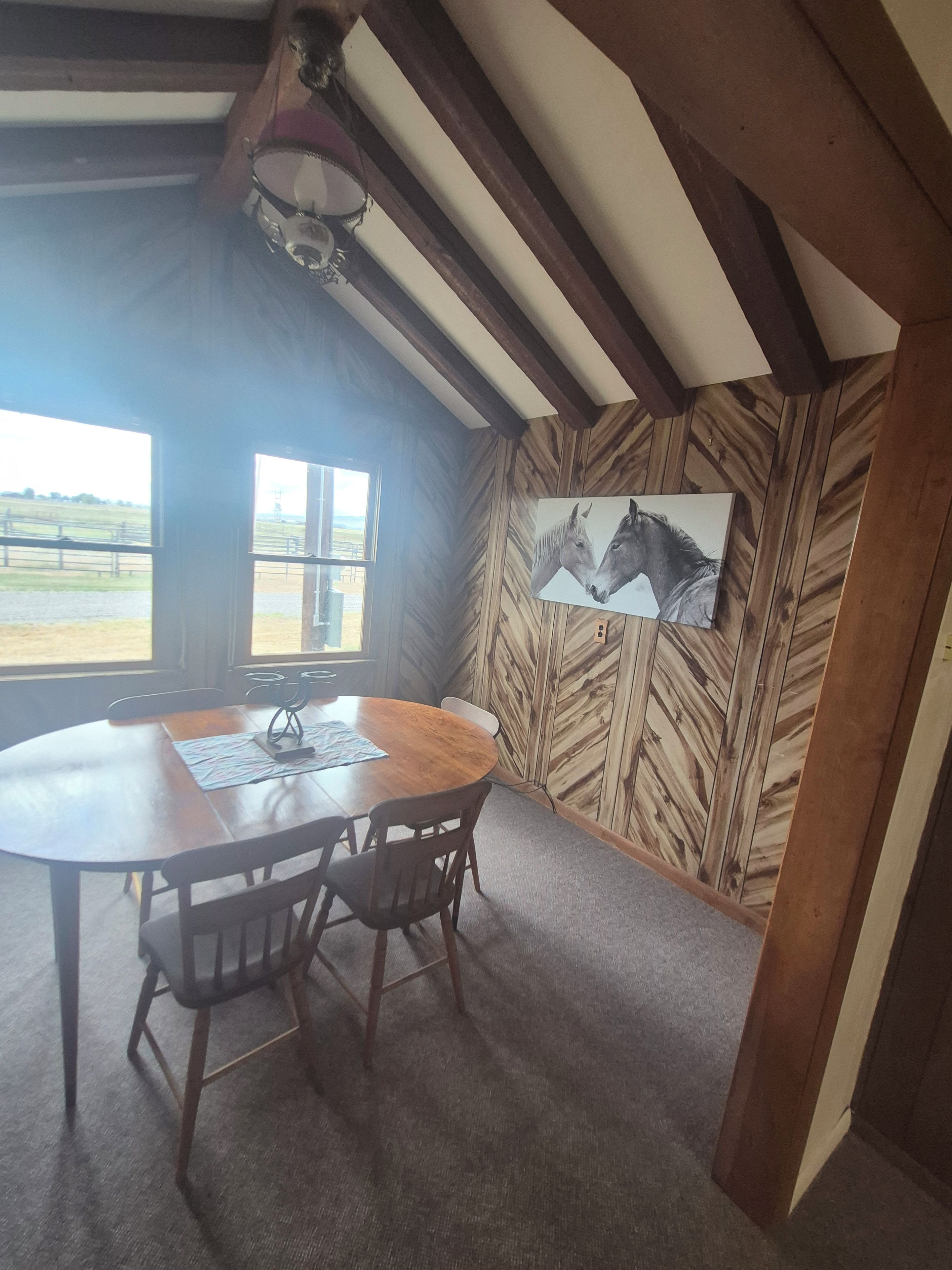 Un coin repas avec une table et des chaises en bois, un tableau représentant des chevaux accroché au mur et une vue sur l’extérieur par une fenêtre.
