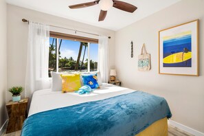 A bedroom with a bed, a ceiling fan, a window with curtains, a beach-themed painting, and a small plant on a nightstand. - Kona Reef#a1: Sand & Surf Beachfront Corner! (Kailua-Kona)