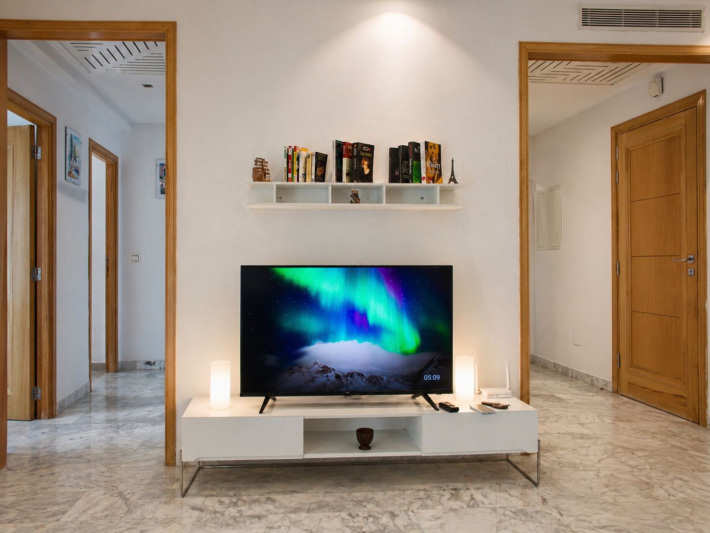 Uma televisão sobre um suporte branco em um corredor, com uma prateleira acima contendo livros e objetos decorativos.