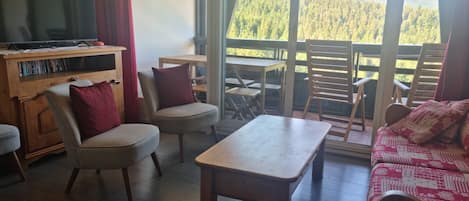 A living room with a TV, seating, a table, and a balcony with a mountain view.