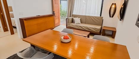 A modern living room with a wooden dining table, two grey chairs, a sofa, and a hanging pendant light.