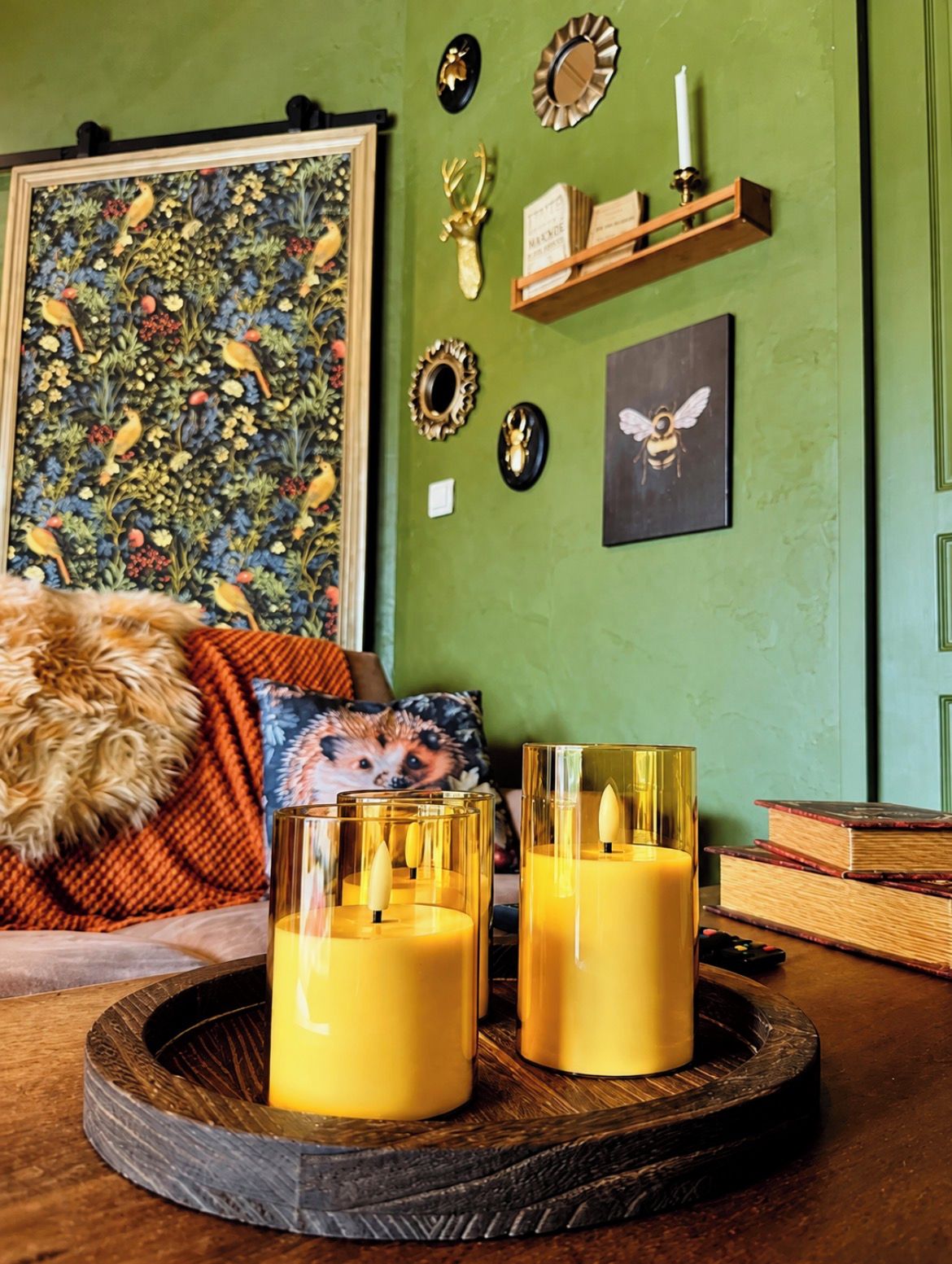Une pièce avec un mur vert, un tableau floral encadré, une étagère avec des objets décoratifs et deux bougies allumées sur un plateau.