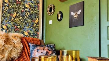 Une pièce avec un mur vert, un tableau floral encadré, une étagère avec des objets décoratifs et deux bougies allumées sur un plateau.