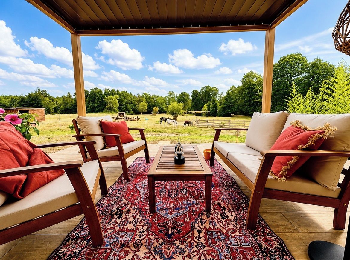 Un espace extérieur aménagé avec une pergola en bois, un tapis à motifs et une vue sur une ferme où paissent des animaux.