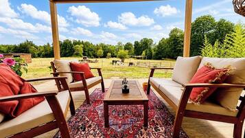 Un espace extérieur aménagé avec une pergola en bois, un tapis à motifs et une vue sur une ferme où paissent des animaux.