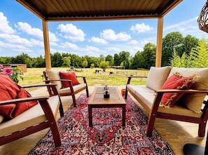 Un área de estar al aire libre con una pérgola de madera, un tapete estampado y vistas a una granja con animales pastando.