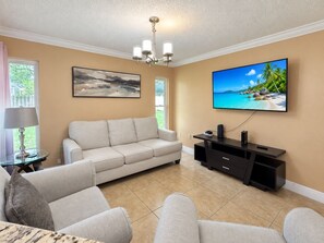 A living room with a beige sofa, a flat-screen TV, a ceiling fan, and a painting on the wall. - Pink Palace - Stylish Sarasota Getaway with Spacious Rooms & Smart TVs (Sarasota)