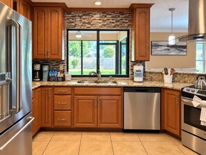 A modern kitchen with wooden cabinets, stainless steel appliances, a coffee bar, and a view of the backyard. - Pink Palace - Stylish Sarasota Getaway with Spacious Rooms & Smart TVs (Sarasota)