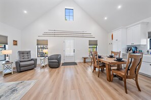A spacious loft with a high ceiling, a dining area with wooden chairs, a kitchen with white cabinets, and a large window providing natural light. - Pinecraft Pearl | Heart of Pinecraft + Rocking Chairs (Sarasota)