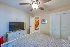 Une chambre à coucher avec un lit, un téléviseur posé sur une commode, un ventilateur de plafond et une porte donnant sur un escalier.