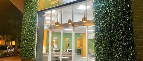 Entrada a un hostal con una pared verde y frondosa, un cartel que dice 'UWANG HOSTEL UP' y un interior visible con asientos y una mesa.