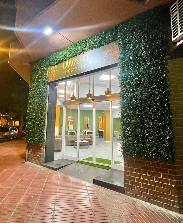 A hostel entrance with a green leafy wall, a sign reading 'UWANG HOSTEL UP,' and a visible interior with seating and a table.