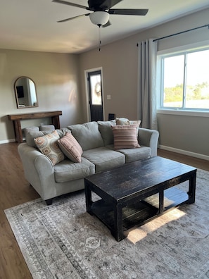 A living room with a sectional sofa, a coffee table, a ceiling fan, and a large window with curtains.
