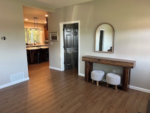 A hallway with wooden flooring, a dark door, a console table with two stools, and a mirror on the wall.
