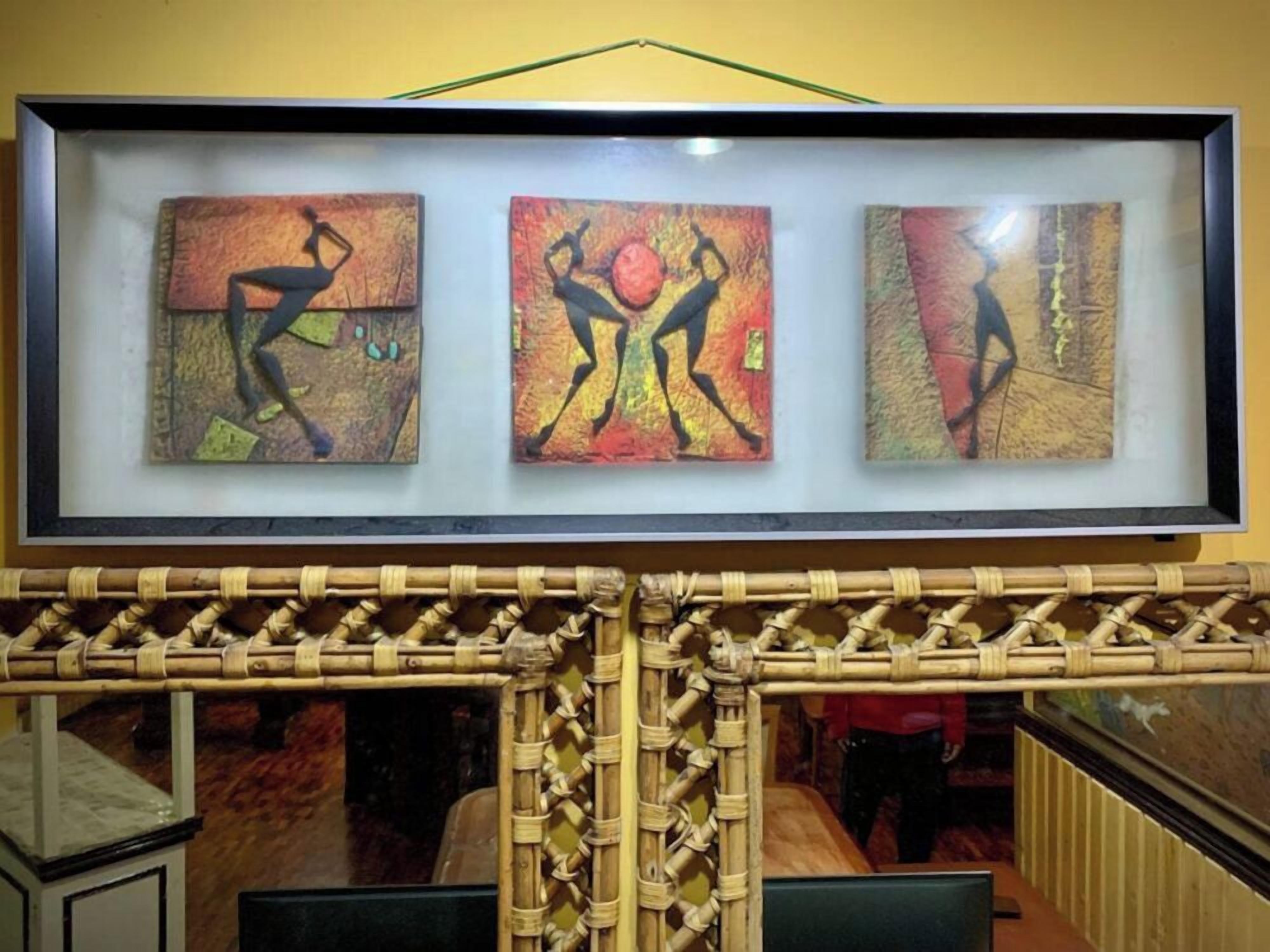 A framed artwork with three panels depicting abstract figures in dynamic poses, displayed above a woven bamboo structure.