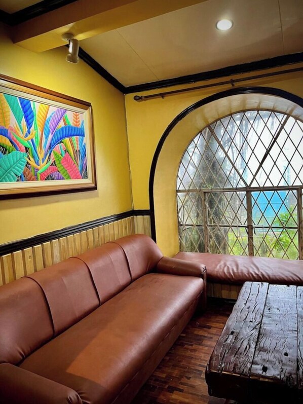 A room with a brown leather sofa, a wooden coffee table, a colorful abstract painting, and a large arched window with a view.