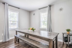 Ein Essbereich mit einem Holztisch und einer Bank, zwei mit Vorhängen versehenen Fenstern und einem kleinen Wanddekor.