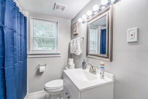 A bathroom with a white sink, a toilet, a mirror with lights, and a blue shower curtain. - Mins to Airport & Dining- Wonderful in Whitehaven (Memphis)