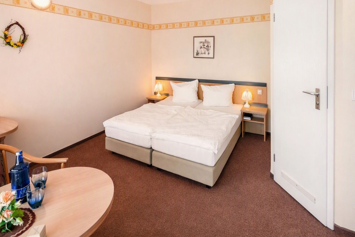 Una habitación de hotel con una cama doble, dos mesitas de noche con lámparas, una pequeña mesa redonda con una botella y un vaso, y un adorno en la pared.