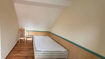 Una habitación pequeña y estrecha con techo inclinado, una cama individual, una silla y una mesita.