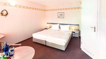 Quarto de hotel com cama de casal, mesas de cabeceira com luminárias, uma mesinha redonda com garrafa e copos, e um quadro pendurado na parede.