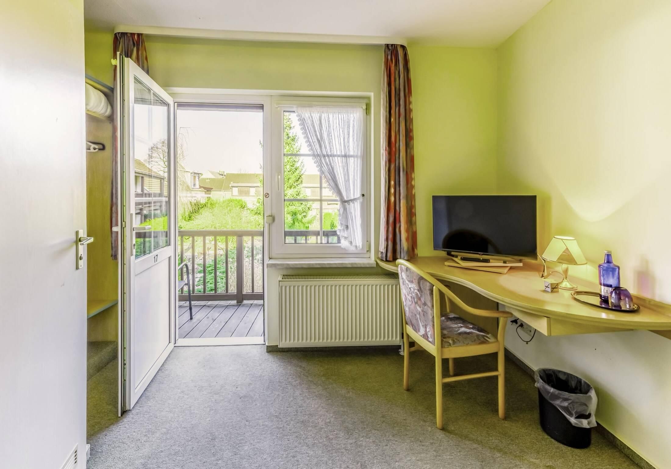 Een kamer met een bureau, een televisie, een radiator, een stoel, een raam met gordijnen en een deur naar een buitenruimte.