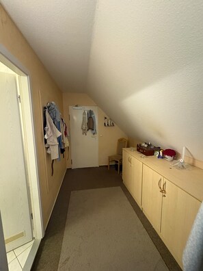 Un pasillo estrecho con una puerta a la izquierda, un perchero con ropa colgada, una silla pequeña y una zona de kitchenette con armarios y una encimera.