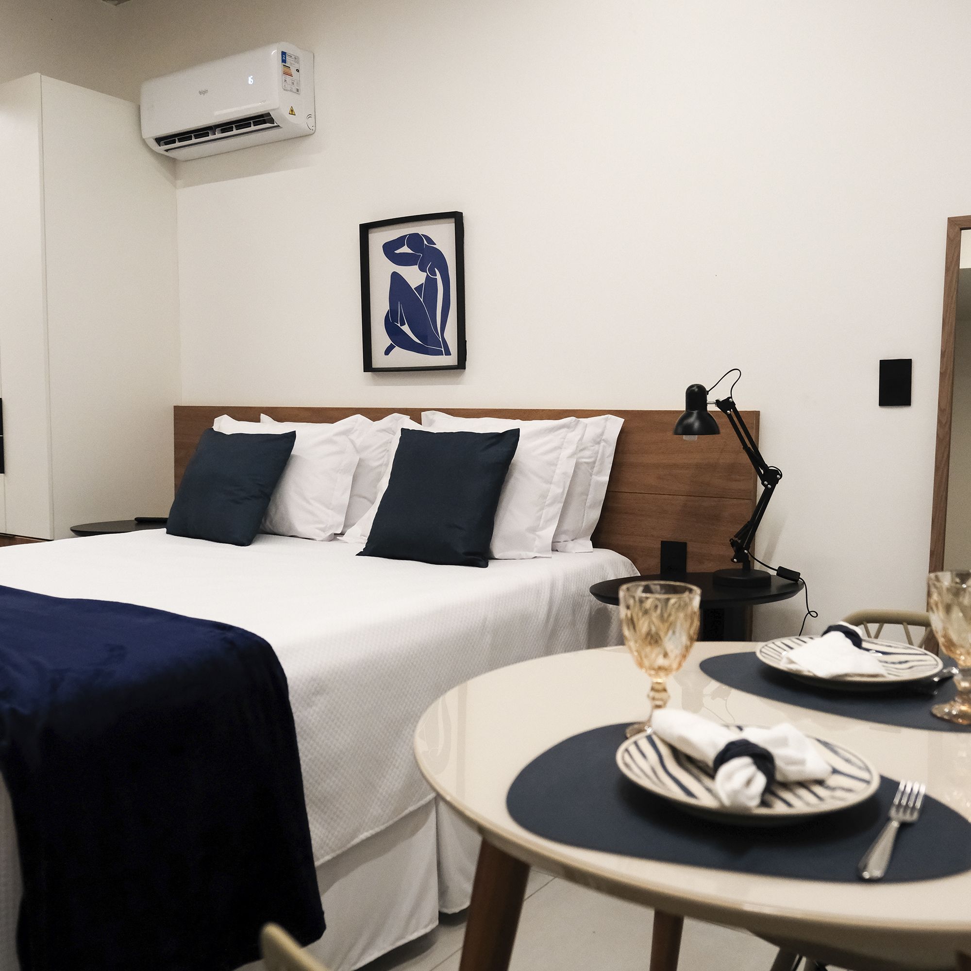 A hotel room with a bed, a bedside table with a lamp, a framed artwork on the wall, and a dining table set with plates and glasses.