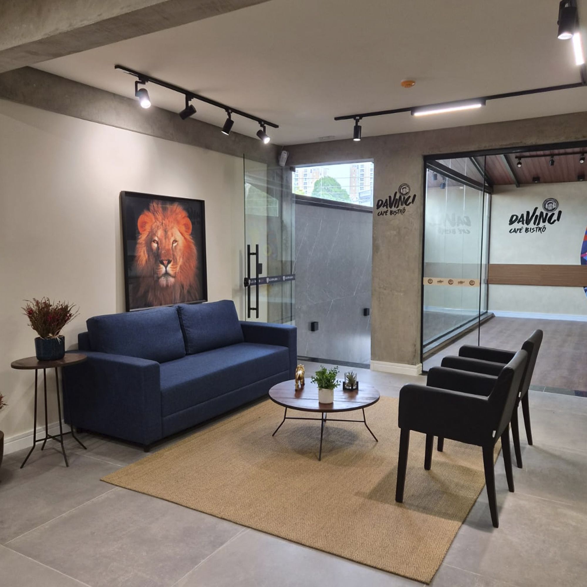 Un interior moderno con un sofá azul, una mesa de centro y sillas negras. Hay una pared de vidrio con vista a la ciudad y un letrero que dice 'Davinci Cafe Bistro'.