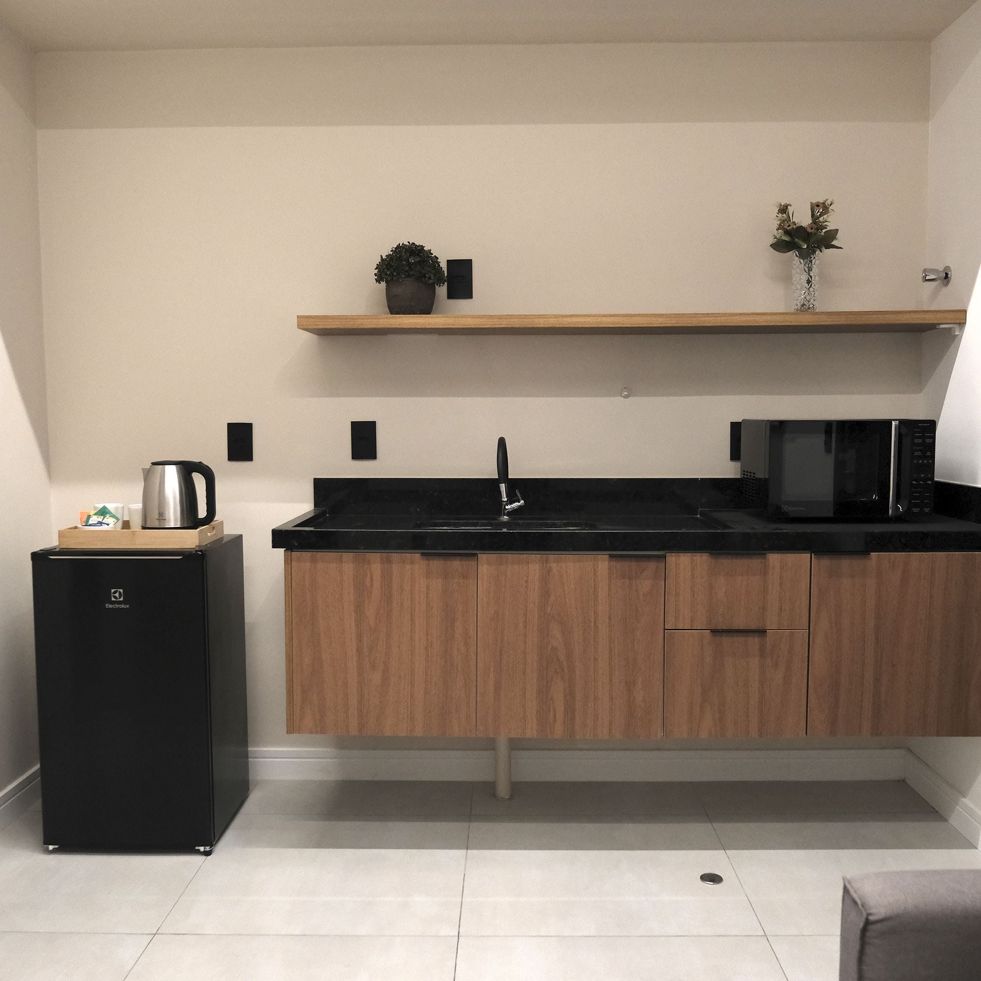 A compact kitchen with a black refrigerator, wooden cabinets, a black countertop, a microwave, a kettle, and a small shelf with a plant.