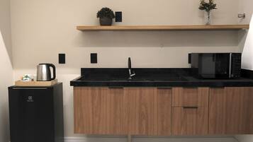 A compact kitchen with a black refrigerator, wooden cabinets, a black countertop, a microwave, a kettle, and a small shelf with a plant.