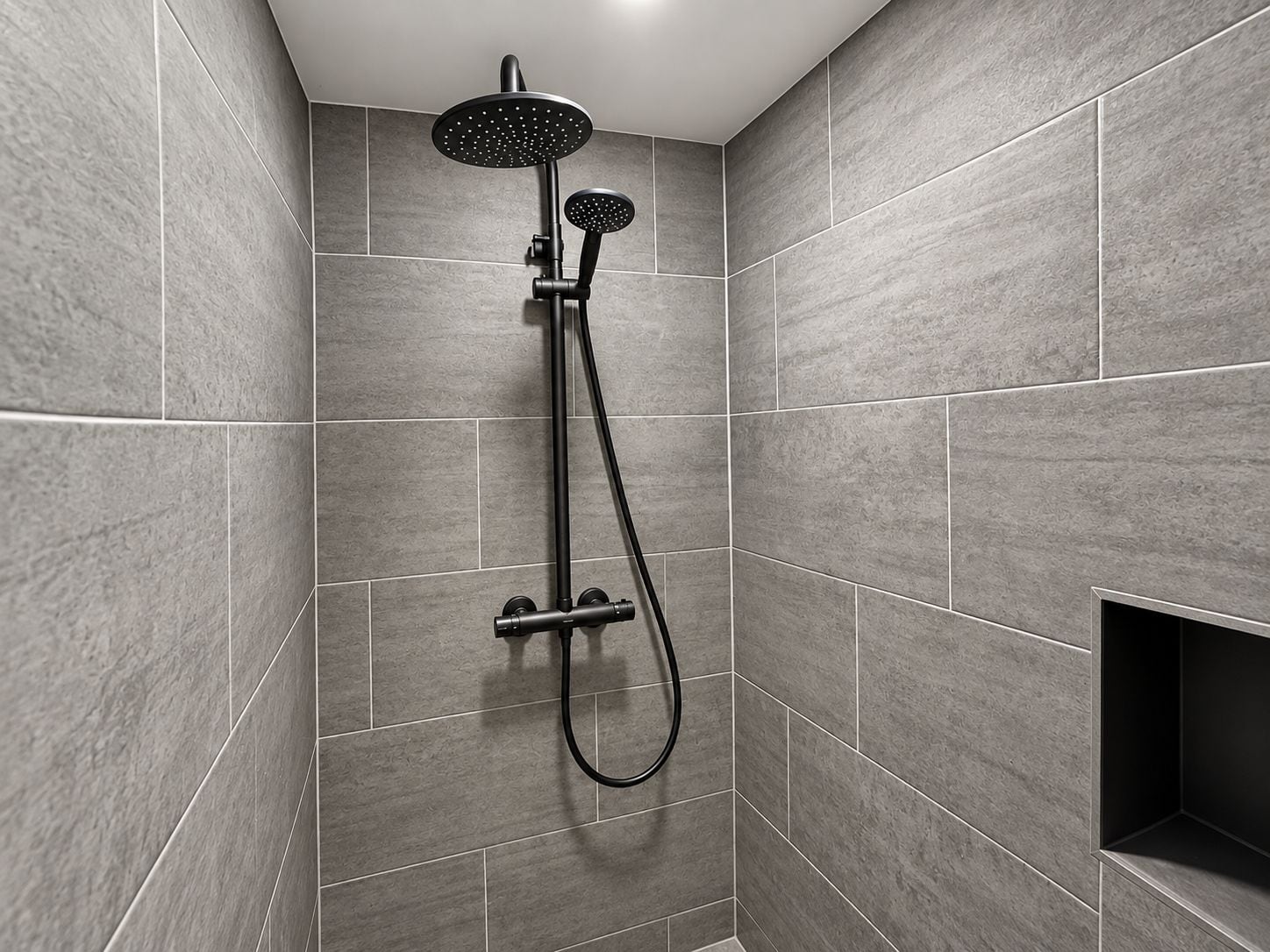 Eine moderne Dusche mit Handbrause und fest installierter Duschbrause, beide mit schwarzen Armaturen, in einer Ecke eines Raumes mit grauen Fliesenwänden.