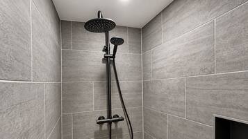 Eine moderne Dusche mit Handbrause und fest installierter Duschbrause, beide mit schwarzen Armaturen, in einer Ecke eines Raumes mit grauen Fliesenwänden.