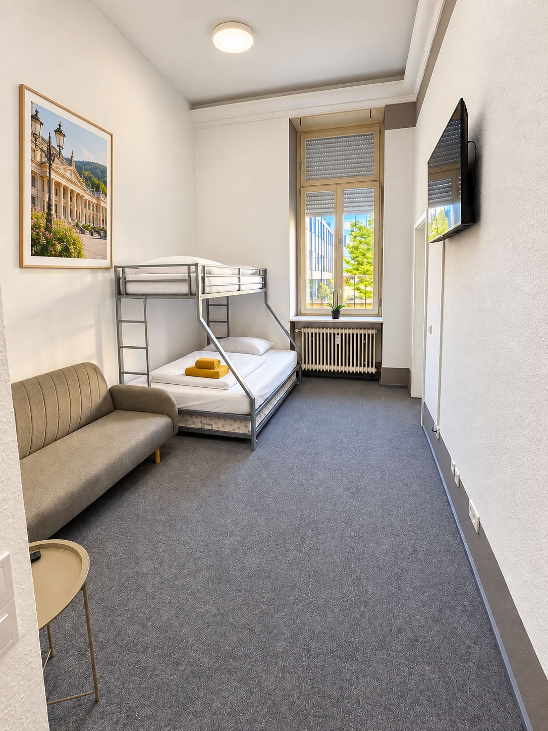 Une pièce compacte comprenant des lits superposés, un canapé, une petite table, un radiateur, une fenêtre avec des stores et un téléviseur fixé au mur.