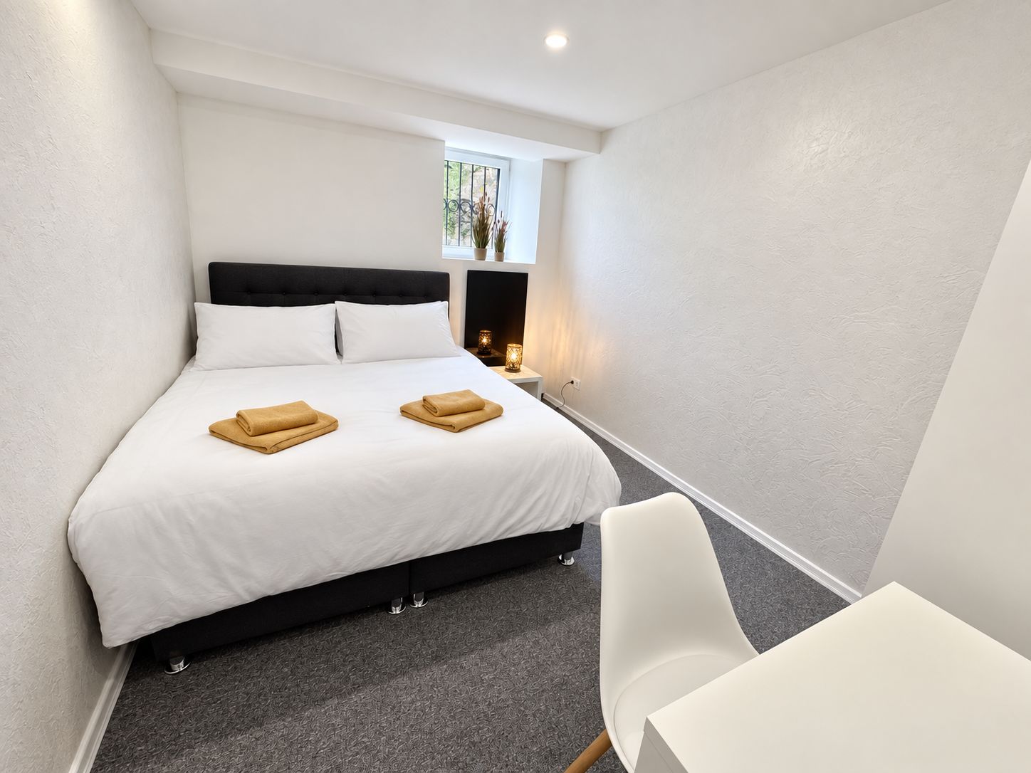 A hotel room with a bed, a bedside table with a lamp, a chair, and a small window.