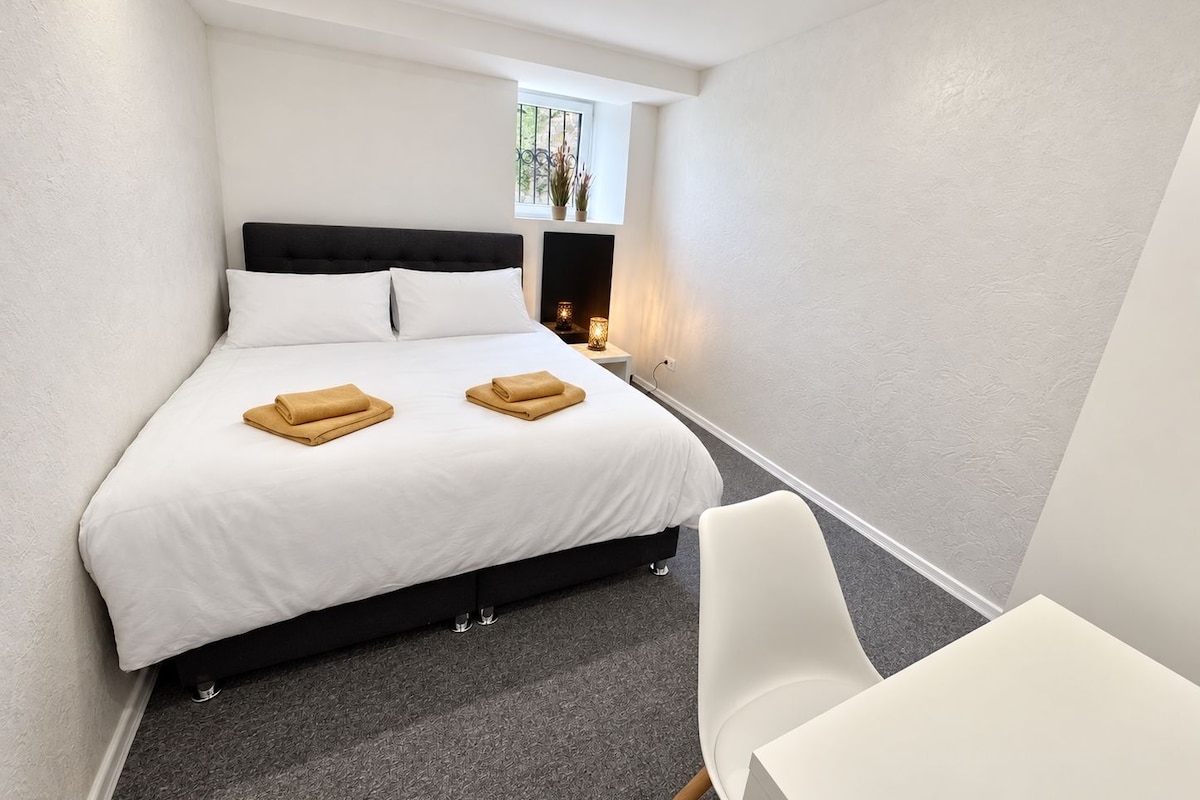 A hotel room with a bed, a bedside table with a lamp, a chair, and a small window.