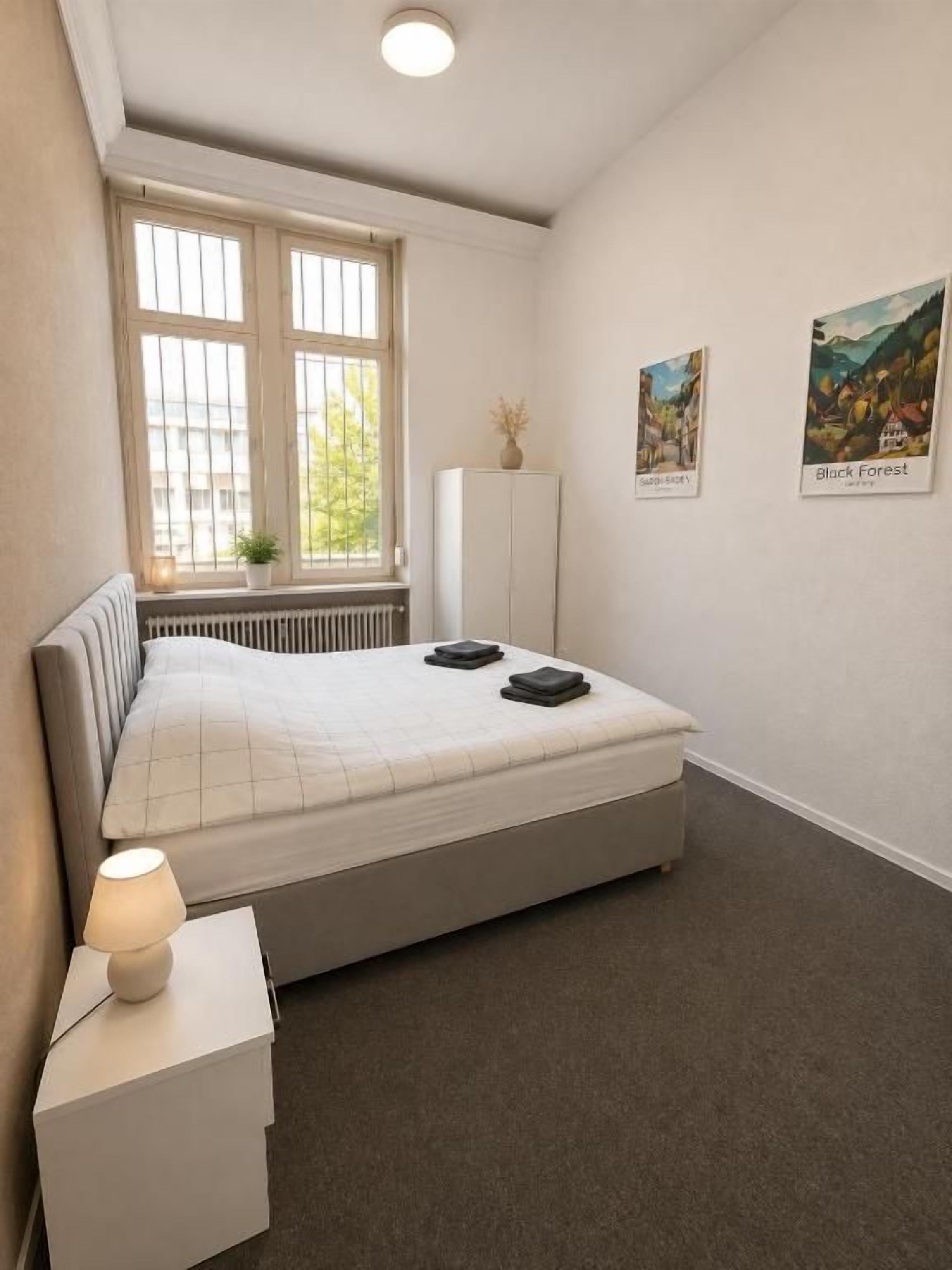 Ein Schlafzimmer mit einem Bett, Nachttisch, Lampe, Heizkörper, Fenster und zwei Wandbildern.