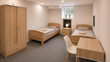 Une chambre avec deux lits simples, une armoire en bois, un bureau avec une chaise et une petite fenêtre ornée de plantes.