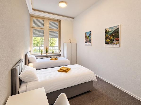 A hotel room with two beds, a window with blinds, a radiator, a wardrobe, and two framed pictures on the wall.